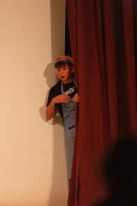 Ein Kind lugt hinter einem dunklen roten Bühnenvorhang hervor. Es trägt ein schwarzes T-Shirt mit weißem Levi's-Logo, darüber eine blau-weiß karierte Schürze und ein buntes Stirnband. Der Blick des Kindes ist nach links gerichtet, der Mund leicht geöffnet. Links im Bild ist eine helle Wand zu sehen, auf die der Schatten des Kindes fällt. Rechts dominiert der Vorhang.