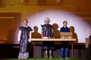 Drei Kinder stehen auf einer Bühne vor einem hellbraunen, holzgetäfelten Hintergrund, der eine formale Halle mit einem goldenen Wappen in der Mitte darstellt. Links steht ein Mädchen in weißem Hemd, blauem Rock und einem schwarzen Müllsack als Robe. Daneben steht ein Junge mit weißer Lockenperücke und ebenfalls einem schwarzen Müllsack über seinem grünen Shirt. Rechts steht ein Junge in blauem Poloshirt und Jeans. Vor ihnen befindet sich ein Holztisch, auf dem ein offenes Buch und ein beiges, kassenähnliches Gerät liegen. Starkes Licht von oben wirft lange, deutliche Schatten der Kinder auf die helle Rückwand. Der Boden ist mit grünem Kunstrasen ausgelegt.