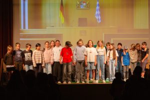 Eine Gruppe von etwa 17 Kindern und Jugendlichen steht in einer Reihe auf einer Bühne. Sie tragen Freizeitkleidung wie T-Shirts, Hoodies und Jeans; einige halten Händchen. Im Hintergrund ist eine Projektion zu sehen, die einen holzvertäfelten, formellen Raum mit Tischen und Rednerpulten darstellt, ähnlich einem Gerichtssaal oder Parlament. Auf der linken Seite der Projektion ist eine deutsche Flagge (Schwarz-Rot-Gold) zu sehen, auf der rechten Seite eine bayerische Rautenflagge (blau-weiß kariert). Zwischen den Flaggen ist ein Wappen an der Wand angebracht. Im Vordergrund sind die dunklen Silhouetten von Zuschauern zu erkennen, die der Bühne zugewandt sind.