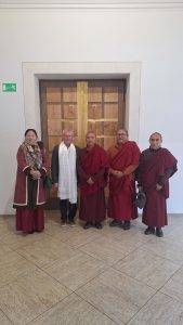 Fünf Personen stehen in einer Innenaufnahme vor einer geschnitzten Holztür. Von links nach rechts: Eine Frau in einem weinroten Mantel mit gemustertem Schal, ein Mann mit Brille und einem weißen Zeremonienschal um den Hals, gefolgt von drei buddhistischen Mönchen in dunkelroten Roben. Die Holztür im Hintergrund weist geschnitzte Felder auf, einen zentralen goldenen Vertikalbalken mit Figuren und der Inschrift "WAS IHR EINEM MEINER GERINGSTEN BRÜDER". Eine der geschnitzten Figuren stellt eine Kreuzigung dar. Links an der weißen Wand befindet sich ein grünes Notausgangsschild. Der Boden ist mit hellbraunen, strukturierten Fliesen ausgelegt.