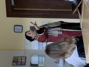 Eine ältere Frau mit dunklem Haar, bekleidet mit einem roten Mantel und einem gemusterten Schal, steht in einem Raum, spricht und gestikuliert mit erhobener rechter Hand. Im Vordergrund, mit dem Rücken zum Betrachter, sitzt eine Person mit blondem Haar an einem Holztisch. Der Hintergrund zeigt eine helle Wand mit einer Pinnwand, auf der Notizzettel und eine kleine Union-Jack-Flagge befestigt sind. Weiter rechts sind eine Wanduhr und eine dunkle Holztür zu sehen. Links im Hintergrund befindet sich ein Waschbeckenbereich mit zwei Handtuchspendern. Auf dem Tisch im Vordergrund liegt ein aufgestelltes Tablet.