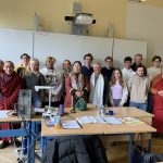Eine Gruppe von etwa 23 Personen steht in einem Klassenzimmer vor einer weißen Tafel und blickt in die Kamera. Mehrere Männer tragen rote oder weinrote Mönchsroben. Die anderen Personen, darunter junge und ältere Erwachsene, sind in Freizeitkleidung gekleidet; ein älterer Mann trägt einen weißen Schal und eine ältere Frau einen gemusterten Schal. Auf der Tafel stehen die Zahlen '7.10.25'. Über der Tafel hängt ein Beamer, und oben links an der Wand ist ein hölzernes Kruzifix zu sehen. Im Vordergrund stehen Holztische mit blauen Beinen, beladen mit Papieren, einer orangefarbenen Getränkeflasche und Computerzubehör.