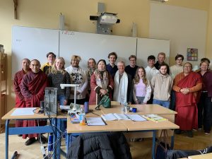 Eine Gruppe von etwa 23 Personen steht in einem Klassenzimmer vor einer weißen Tafel und blickt in die Kamera. Mehrere Männer tragen rote oder weinrote Mönchsroben. Die anderen Personen, darunter junge und ältere Erwachsene, sind in Freizeitkleidung gekleidet; ein älterer Mann trägt einen weißen Schal und eine ältere Frau einen gemusterten Schal. Auf der Tafel stehen die Zahlen '7.10.25'. Über der Tafel hängt ein Beamer, und oben links an der Wand ist ein hölzernes Kruzifix zu sehen. Im Vordergrund stehen Holztische mit blauen Beinen, beladen mit Papieren, einer orangefarbenen Getränkeflasche und Computerzubehör.