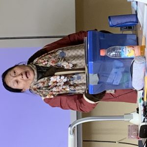 Eine Frau mittleren bis höheren Alters mit dunklen Haaren steht hinter einem Tisch und spricht, der Mund ist leicht geöffnet. Sie trägt einen rotbraunen Mantel und einen beigen Schal mit floralem Muster und einem wiederholten Markenlogo. Auf dem Tisch vor ihr befinden sich ein blauer Aufbewahrungsbehälter, eine Flasche mit einem pfirsichfarbenen Getränk, eine graue Stiftetasche, ein Laptop, eine Schreibtischlampe, ein kleiner Tiegel mit rosa Inhalt, ein elektronisches Gerät und diverse Kabel. Im Hintergrund sind eine große lilafarbene Projektionsfläche und eine helle Wand zu sehen.